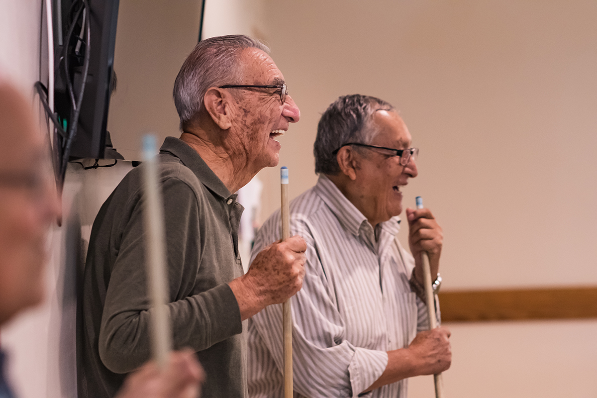 Two older men speaking to each other while holding pool cues.