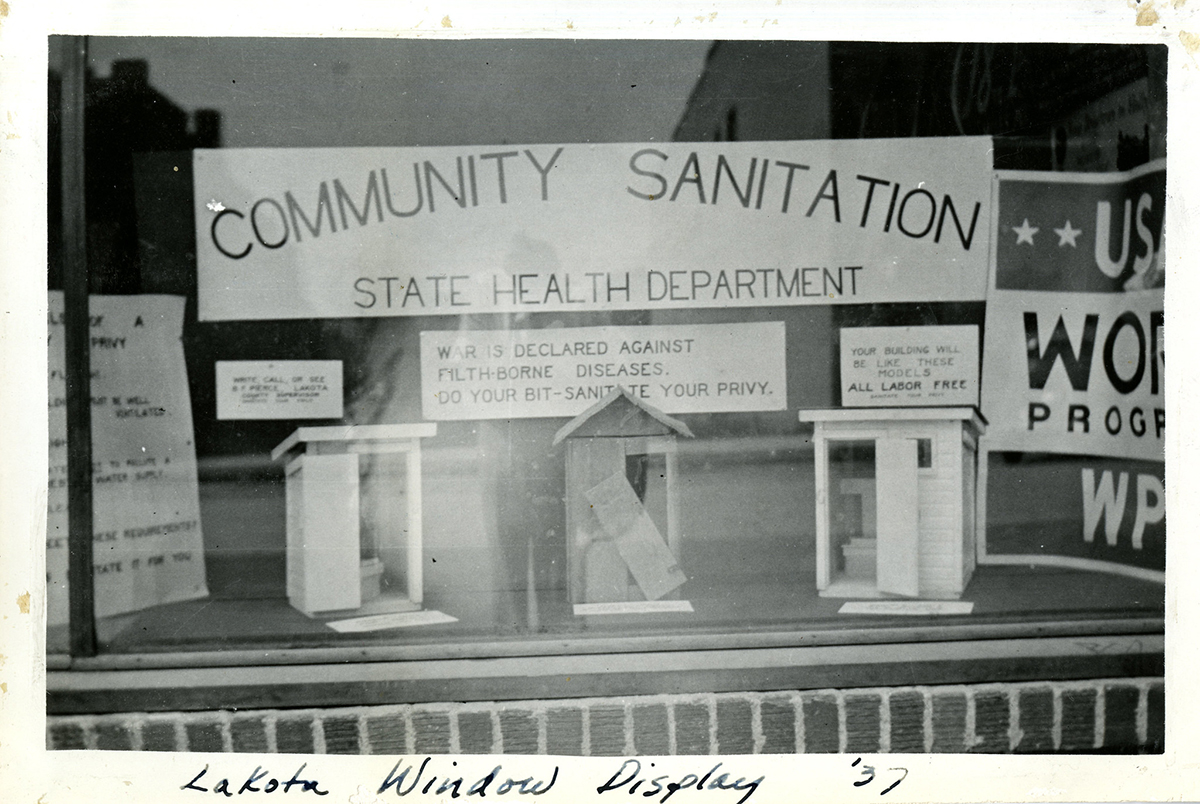 Exterior view of window display showing three model privies under a sign that reads "Community Sanitation."