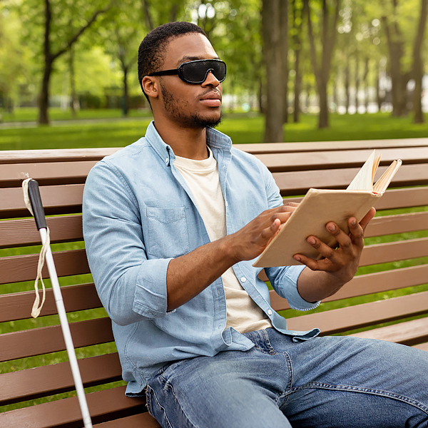 A vision impaired man sitting on a park bench and reading a braille book