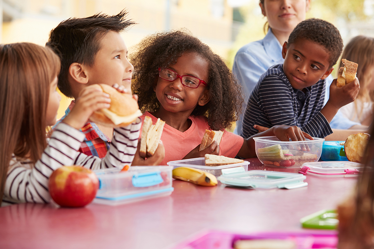 Four children eat together at a table. Three children look with interest at the fourth child.