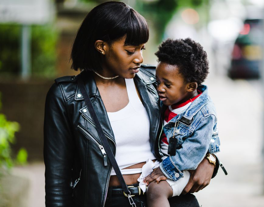 Black woman holding a toddler and walking down a city street.