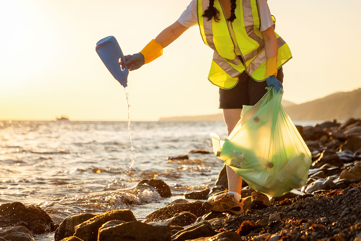 Person in a safety vest cleaning up debris from the ocean and a rocky shore. They are pouring water out of a plastic, liquid-dispensing bottle that was collected. 