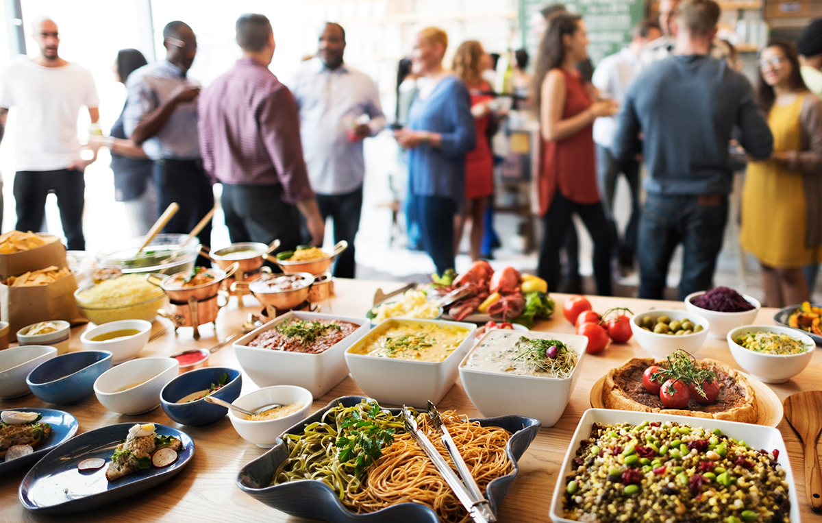 A table displaying a variety of foods with a crowd of people standing around and talking behind it. A table displaying a variety of foods with a crowd of people standing around and talking behind it.