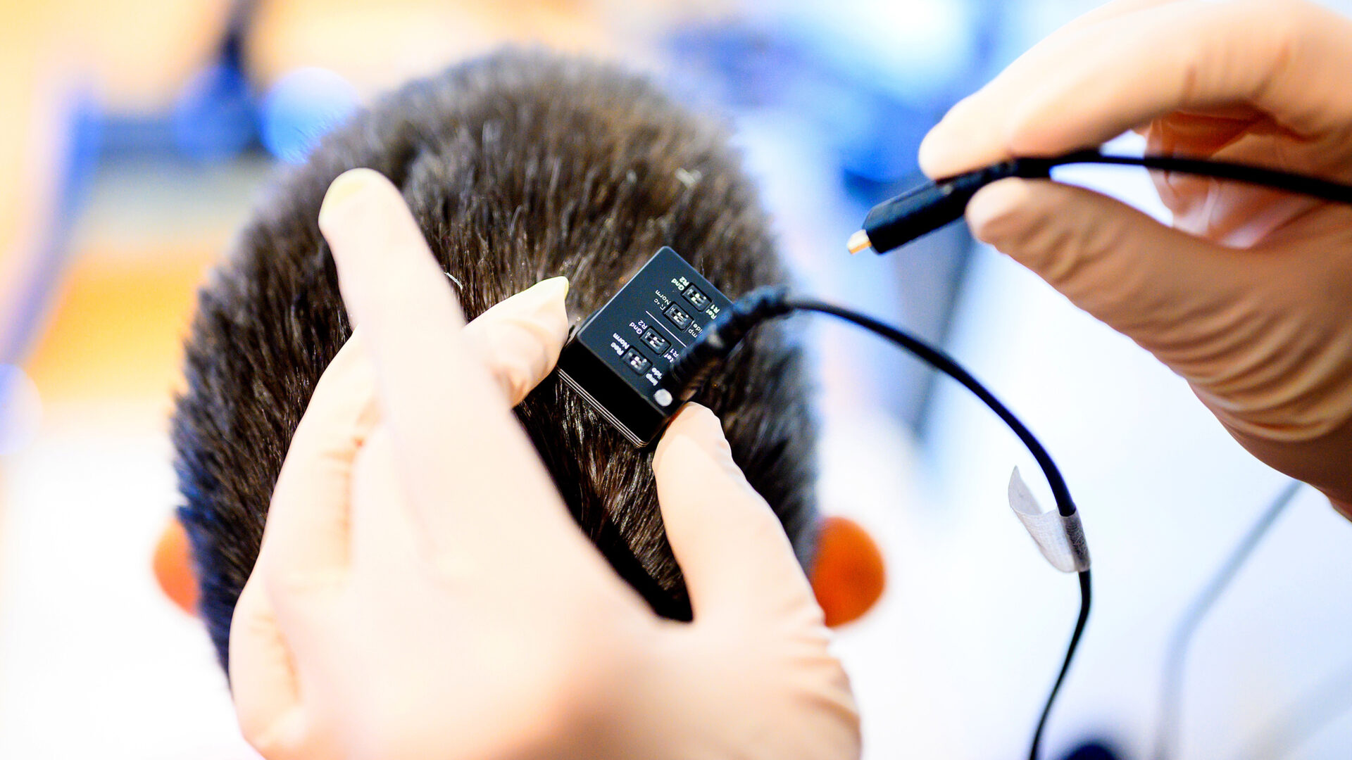 Researcher plugging a cable into a device on a person’s head. Researcher plugging a cable into a device on a person’s head.