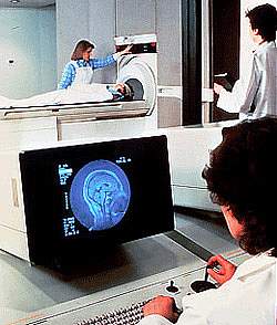 Image of brain on computer screen in foreground, with patient in background lying on an MRI machine Image of brain on computer screen in foreground, with patient in background lying on an MRI machine