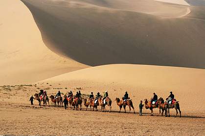 Photo of people riding camels past sand dunes Photo of people riding camels past sand dunes