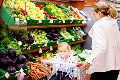 A woman choosing vegetables at a grocery with a baby.