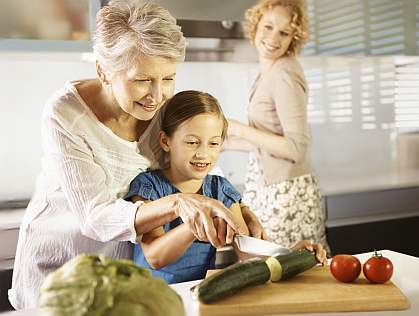 A grandmother helping her granddaughter slice a cucumber as her mother looks on.