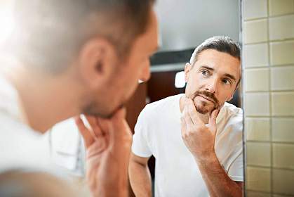 Mature man looking in bathroom mirror at the gray hair in his beard Mature man looking in bathroom mirror at the gray hair in his beard