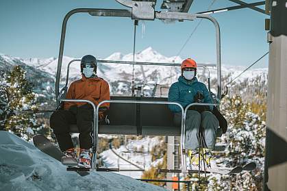 Two skiers with face masks on each side of a chair lift Two skiers with face masks on each side of a chair lift