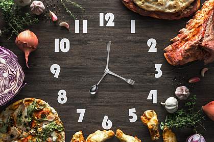 Black wooden clock with utensils as hands and surrounded by food Black wooden clock with utensils as hands and surrounded by food