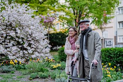 Senior man with a walker and a younger woman strolling outdoors in an urban park.