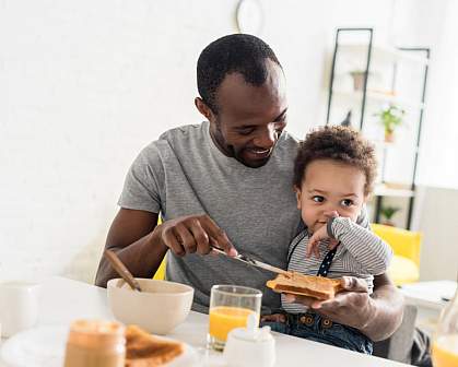 Father with young son on his lap spreading peanut butter on toast.