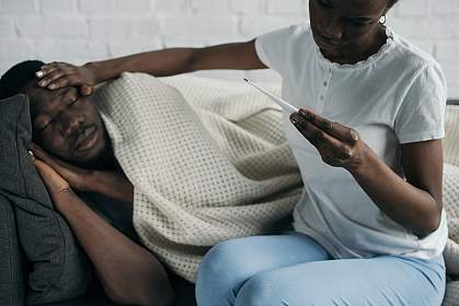 A young woman reading a thermometer and checking the forehead of a sick young man on a couch. A young woman reading a thermometer and checking the forehead of a sick young man on a couch.