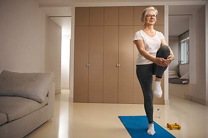 Older woman standing on one leg on a mat in the living room. Older woman standing on one leg on a mat in the living room.