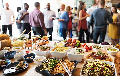 A table displaying a variety of foods with a crowd of people standing around and talking behind it. A table displaying a variety of foods with a crowd of people standing around and talking behind it.