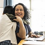 Woman sitting at home at a table working on her laptop and smiling while her pre-teen son hugs her.