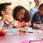 Four children eat together at a table. Three children look with interest at the fourth child.