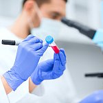 Laboratory scene with scientists’ hands labelling a test tube while another researcher in the background looks through a microscope.