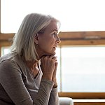 Side view of thoughtful middle-aged mature woman sitting on a couch at home in front of a window.